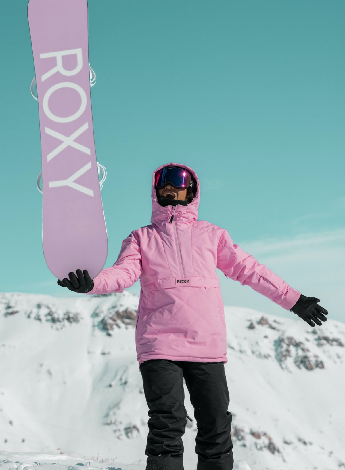 a snowboarder in black pants and pink Roxy jacket balancing a pink Roxy snowboard on one hand