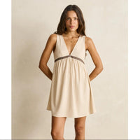 A woman modeling a sleeveless beige mini dress with a deep V-neckline and a dark patterned waistband, set against a neutral textured beige background.