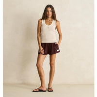 A woman stands in a studio wearing a ribbed cream tank top and dark burgundy athletic shorts, with black sandals, against a warm beige textured wall.