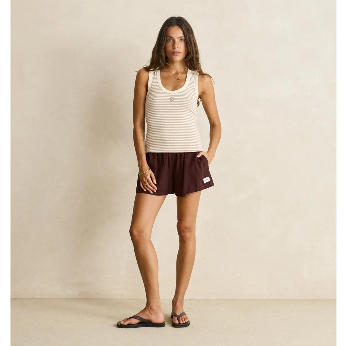 A woman stands in a studio wearing a ribbed cream tank top and dark burgundy athletic shorts, with black sandals, against a warm beige textured wall.