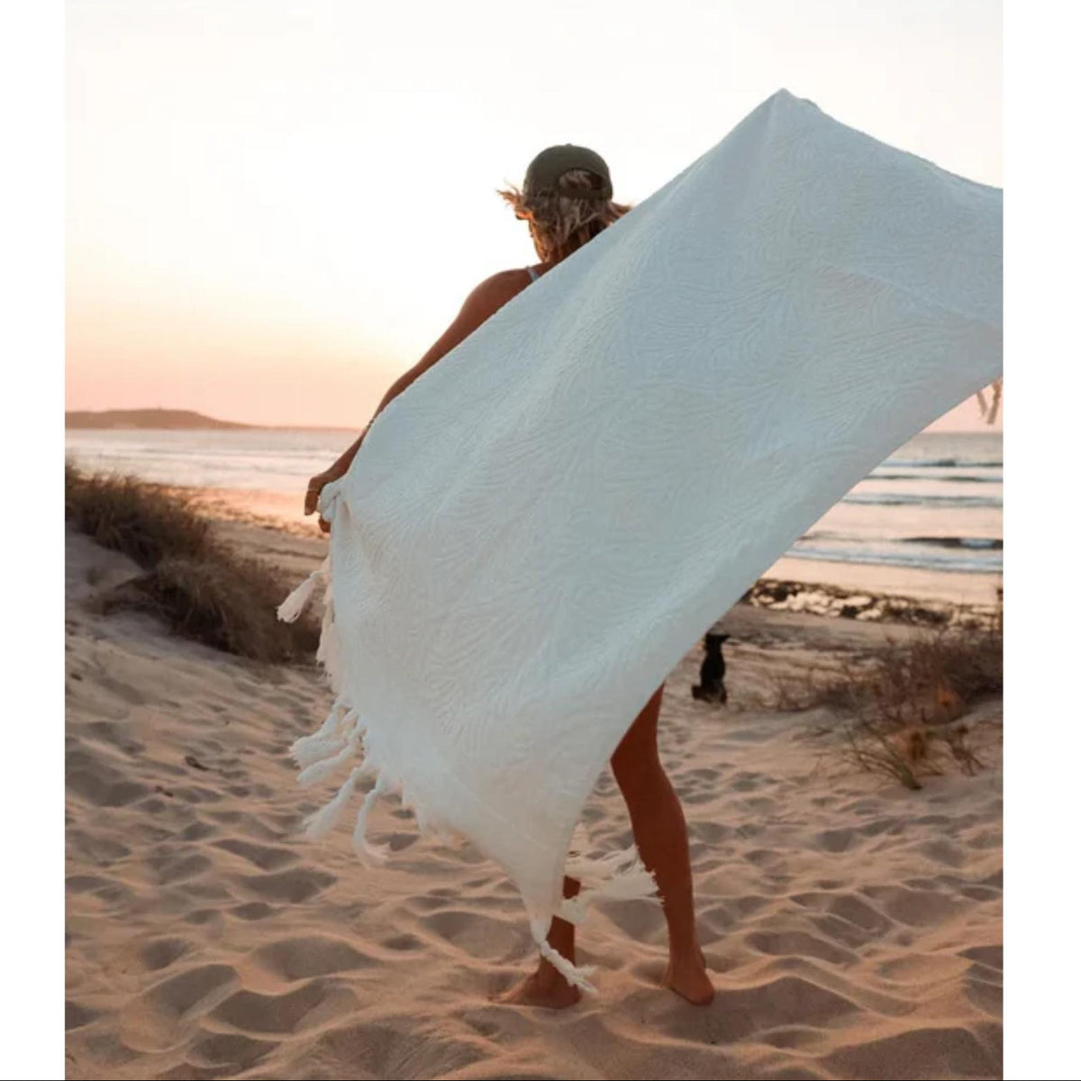 A woman on a sandy beach at sunset holds up a large white woven towel or blanket with tasseled edges, with the ocean and sky in the background.