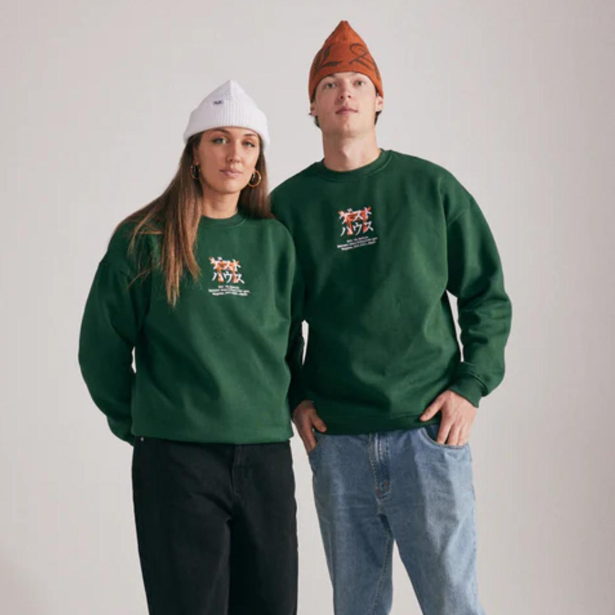 Two people wearing green sweatshirts and beanies on a plain background