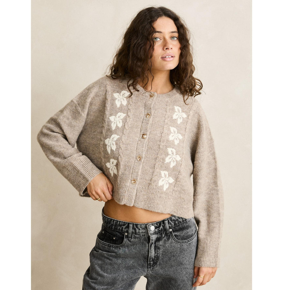 Woman wearing a beige cardigan with floral patterns and gray jeans against a beige background