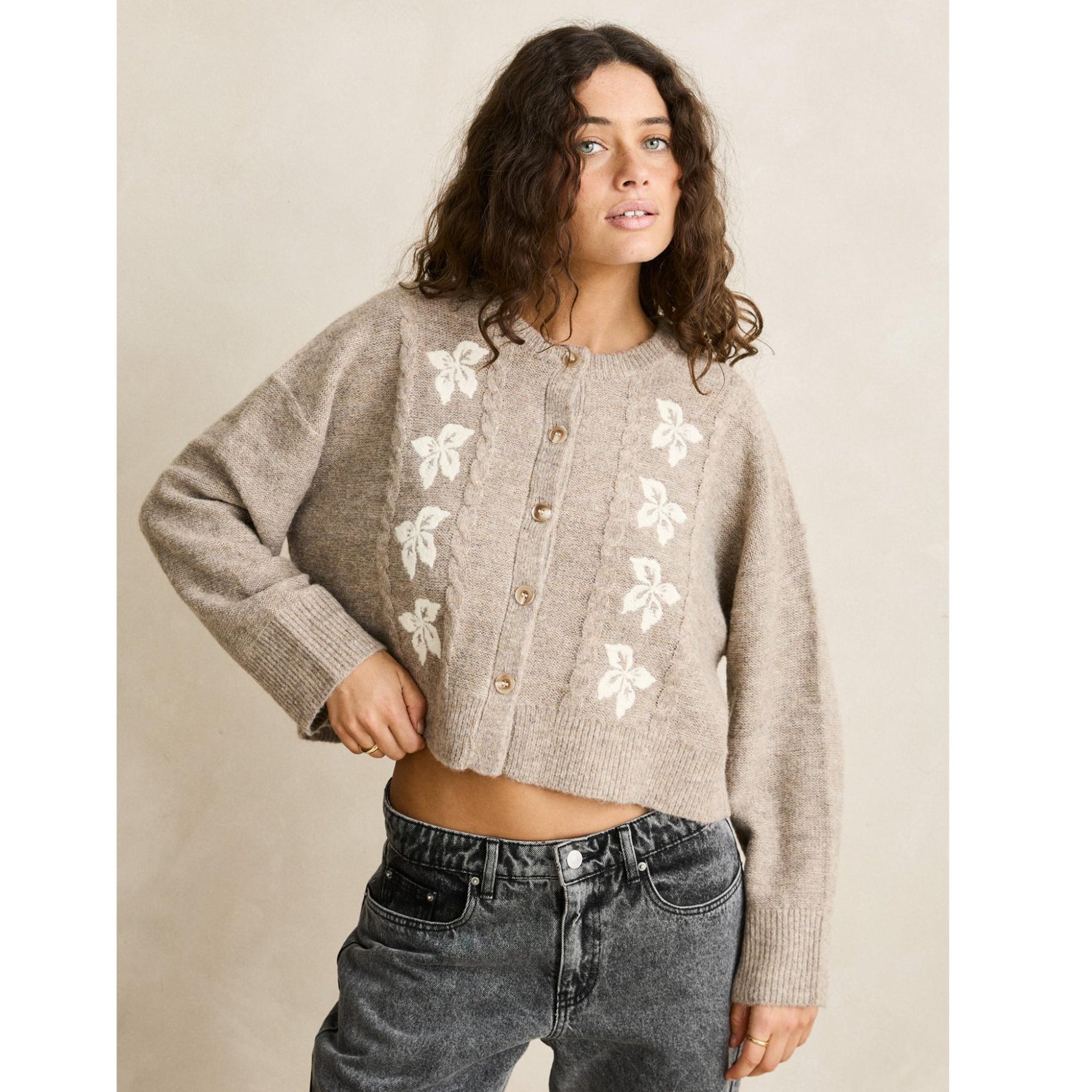 Woman wearing a beige cardigan with floral patterns and gray jeans against a beige background